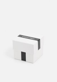 Witte doos met een zwarte streep en het "Calvin Klein" logo aan de voorkant. Vierkante vorm, gladde textuur, minimalistisch ontwerp.