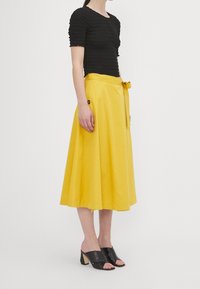 Top in smock nero con maniche corte abbinato a una gonna gialla a taglio A lunga fino al ginocchio con cintura a fiocco e bottone decorativo marrone. Tacchi aperti neri.