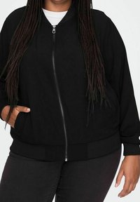 Giacca bomber nera con zip, vestibilità morbida, tessuto leggero, polsini e orlo a coste, e tasche laterali. Texture liscia con dettagli minimi.