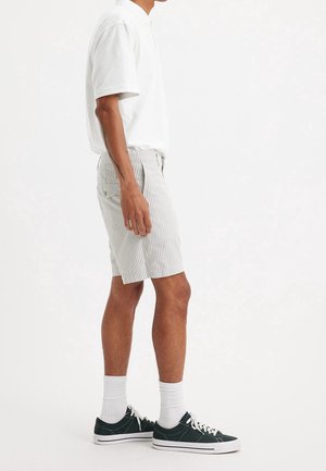 Weißes Polohemd mit kurzen Ärmeln kombiniert mit hellgrau-weiß gestreiften Shorts. Abgerundet mit grünen Sneakers und weißen Knöchelsocken.