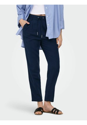 Femme portant un pantalon à cordon de serrage bleu marine, des sandales plates noires, un haut blanc court et une chemise à rayures bleues boutonnée avec les manches retroussées.