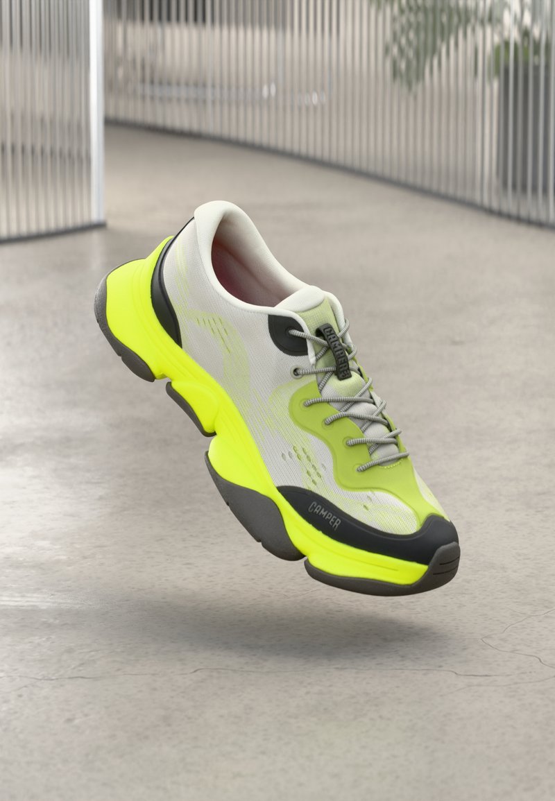 Hvit og neongul treningssko med grå lissesvevende over et polert betonggulv i en moderne, metallisk innendørs setting.