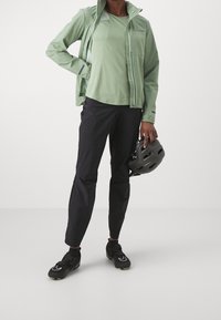 Vaaleanvihreä softshell-takki, samansävyinen vihreä paita, mustat kevyet housut ja mustat urheilukengät. Pitelee harmaata pyöräilykypärää.