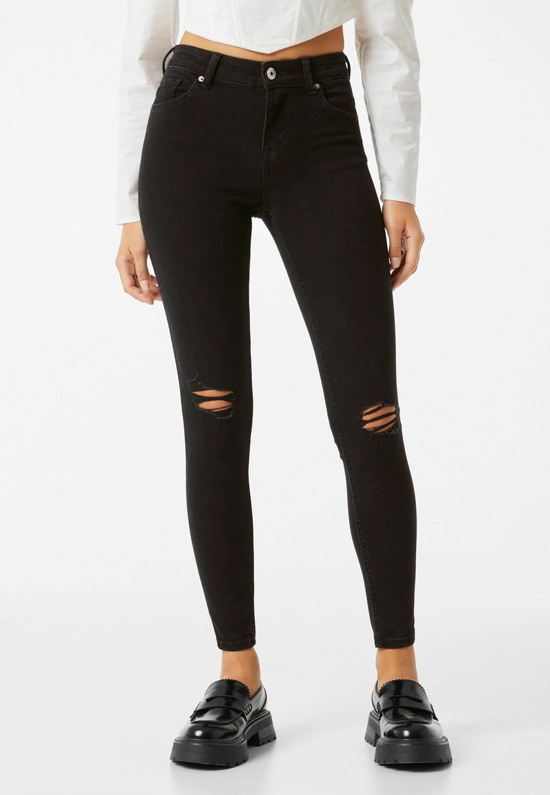 Jeans skinny negros de tiro alto fabricados en denim, con desgastes en las rodillas, cinco bolsillos y un cierre clásico de botón y cremallera.