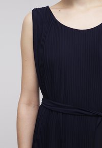 Tummansininen laskostettu mekko pyöreällä kaula-aukolla, olkaimeton malli ja sidottu vyötärötieto. Silkkinen kangas, jossa on pystysuora tekstuuri.