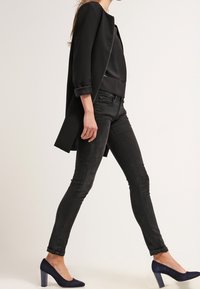 Cappotto nero con una silhouette dritta, abbinato a un top nero e jeans slim-fit. Il modello indossa scarpe con tacco alte di colore blu navy. Texture lisce.