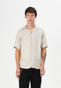 Light beige short-sleeve shirt with a collar, front buttons, and smooth texture, paired with black pants and a silver chain detail.