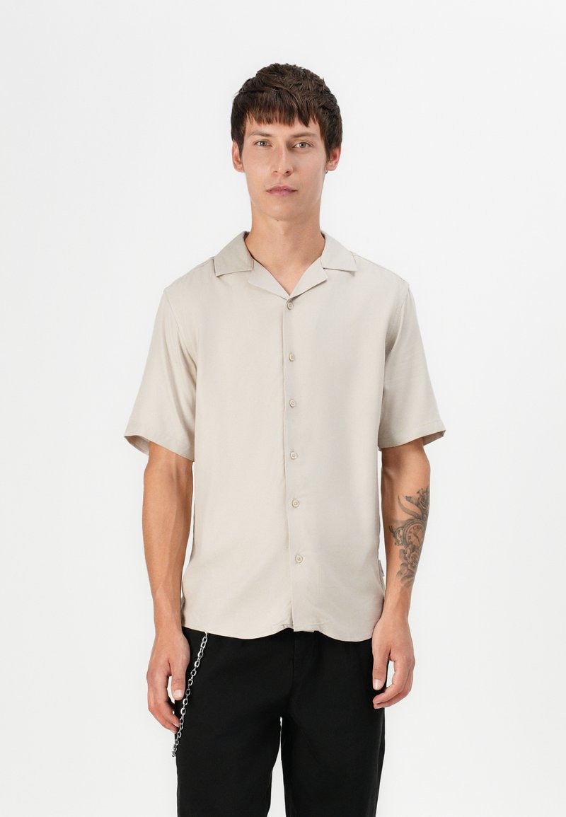 Light beige short-sleeve shirt with a collar, front buttons, and smooth texture, paired with black pants and a silver chain detail.