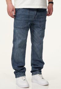 Dunkelblaue Denim-Jeans mit entspannter Passform, ausgestattet mit fünf Taschen und sichtbaren Steppnähten. Kombiniert mit weißen Sneakern.