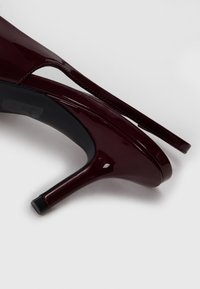 Chaussure à talon aiguille en cuir verni bordeaux avec un bout pointu, une sangle fine et un talon texturé noir. Fini lisse et brillant.