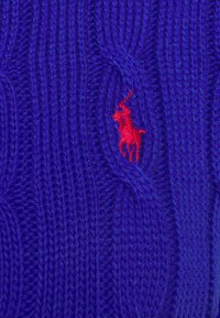 Lila stickad tröja med kabelstruktur. Har en liten röd broderad logotyp av en polo-spelare på framsidan.