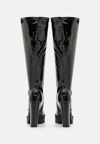 Musta patenttipintainen polven yli yltävä saapas, jossa on kiiltävä pinta, lohkarepohjat ja jämäkkä muotoilu, jossa on sauman yksityiskohta takana.