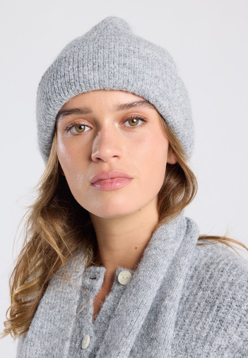 Gray knit beanie features a rounded top and ribbed texture. The model wears a matching gray cardigan with buttons visible.