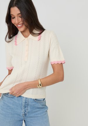 Donna sorridente che indossa un top corto in maglia color crema con bordi smerlati rosa e jeans azzurri, accessoriata con un braccialetto d'oro.