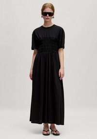 Zwarte maxi-jurk van zachte stof, met korte mouwen en een gerimpelde taille. Model gestyled met zwarte zonnebril en sandalen.