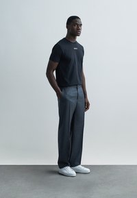 Svart kortärmad t-shirt med "Versace"-logotyp, kombinerad med grå skräddarsydda byxor och vita sneakers, på en enfärgad ljus bakgrund.