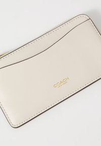 Light beige leather wallet with subtle texture, dark brown stitching, and a gold logo. Compact design with a curved top edge detail.