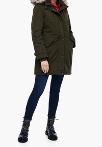 Parka en verde oliva con capucha forrada de piel, botones frontales y dos bolsillos laterales, combinada con jeans ajustados azul oscuro y botas negras con cordones.