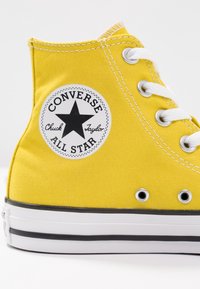 Gul højt snøresko i lærred med hvide snørehuller og snørebånd, med en sort stjerne-logo og sort gummisål med en hvid stribe.