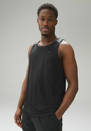 Mann trägt schwarzes ärmelloses Sportshirt und schwarze Shorts, steht mit einer Hand in der Tasche vor einfachem hellem Hintergrund.