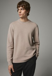 Beżowy sweter dzianinowy z prążkowanym okrągłym dekoltem, długimi rękawami i teksturowanym materiałem. Połączony z ciemnymi spodniami w prążki, modelowany przez mężczyznę.