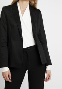 Blazer negro ajustado con una textura suave, solapas con muesca y cierre de un solo botón, combinado con una blusa blanca cruzada y pantalones negros.