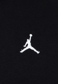 Fekete anyag fehér hímzett Jumpman logóval, amely egy dinamikus pózban lévő sziluettet ábrázol, a fején egy kosárlabdával.