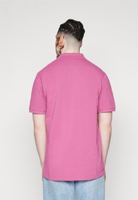Camisa polo de manga corta en algodón rosa sólido. Presenta un cuello clásico y costuras limpias. La vista posterior muestra un ajuste relajado y una textura suave.