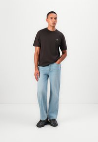 Men's dark brown cotton t-shirt with short sleeves, paired with light blue straight-leg jeans. Notable green logo on the chest. Black footwear.