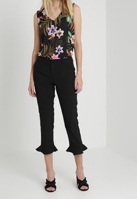 Top senza maniche a fiori con base nera, abbinato a pantaloni neri a vita alta con orli arricciati e tacchi neri aperti.