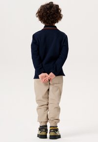 Marineblau strukturiertes Langarmshirt, beige gefesselte Hose, schwarz-graue Schuhe mit gelben Akzenten, Kind steht mit den Händen hinter dem Rücken.