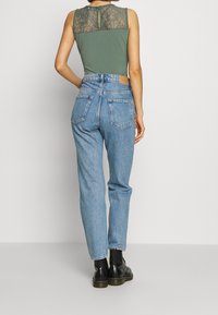 Olivgrön ärmlös topp med spetsdetaljer på baksidan, kombinerad med högmidjade ljusblå denimjeans och svarta ankelstövlar.