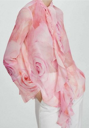Elle porte un chemisier rose transparent avec de grands motifs de roses et un devant à volants, associé à un pantalon blanc, les mains dans les poches.