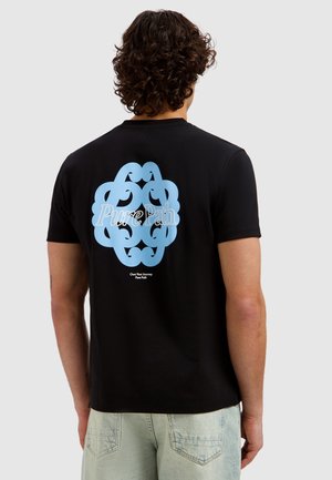 Man met krullend haar, gekleed in een zwart T-shirt met een groot lichtblauw abstract ontwerp en de tekst "Pure Path" op de achterkant, lichte spijkerbroek.
