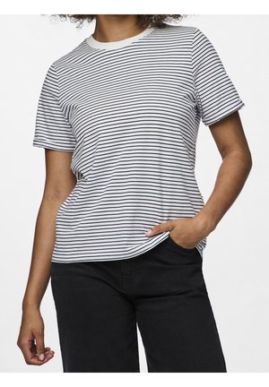 Personne aux cheveux bouclés portant un T-shirt blanc à manches courtes avec de fines rayures horizontales noires et un pantalon noir, main dans la poche.