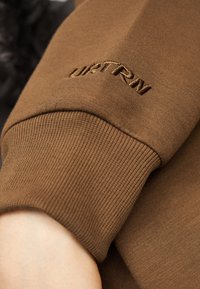Καφέ μανίκι φούτερ με ριμπ μανσέτα, με κεντημένο το "ULTRM" σε λίγο πιο ανοιχτό καφέ νήμα. Ομαλή υφή σε όλο το μήκος.