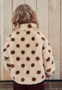 Veste en fausse fourrure beige clair avec de grands pois marron. Elle possède un col montant et une coupe décontractée, associée à un pantalon en velours côtelé bordeaux.