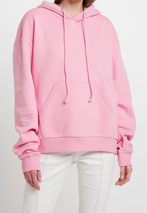 Vrouw draagt een oversized lichtroze hoodie met voorzak en witte broek met zichtbare stiksels.