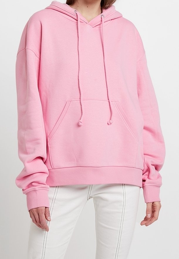 Femme portant un sweat à capuche oversize rose clair avec poche avant et pantalon blanc avec coutures visibles.