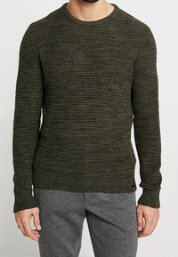 Homme portant un pull en maille texturée vert olive et un pantalon gris, debout devant un fond clair uni.