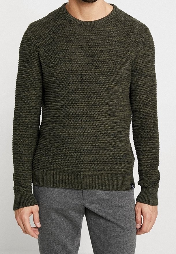 Homme portant un pull en maille texturée vert olive et un pantalon gris, debout devant un fond clair uni.