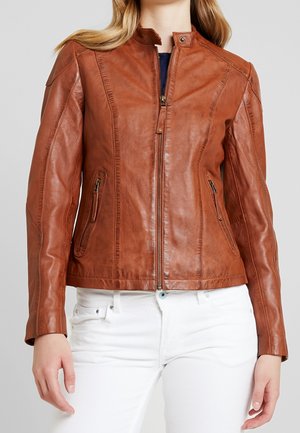 Femme portant une veste en cuir marron ajustée avec des poches zippées et un pantalon blanc, vue des épaules aux hanches.