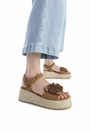 Sandalias de cuña de ante marrón con un acento floral, plataforma envuelta en yute y tira al tobillo. Llevadas con pantalones de mezclilla de pierna ancha en azul claro.