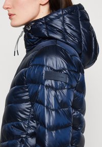 Veste matelassée bleu marine avec capuche, présentant un design matelassé, une texture brillante, un cordon de serrage réglable et un patch logo sur la manche.