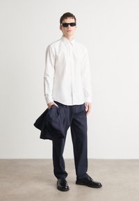 Biely košeľový top, námornícke nohavice, čierne topánky. Model drží námornícky sako, nosí čierne slnečné okuliare a stojí na svetlom pozadí.