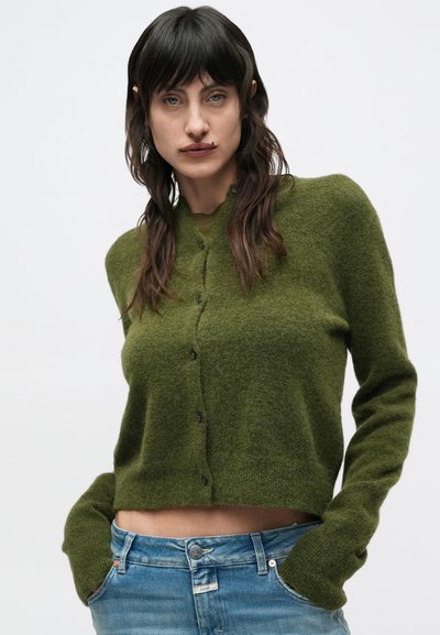 Γυναίκα με μακριά σκούρα μαλλιά, που φοράει πράσινη κοντή ζακέτα και μπλε τζιν, στέκεται με τα χέρια στις τσέπες μπροστά σε απλό λευκό φόντο.