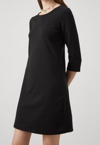Femme portant une robe noire simple arrivant au genou avec des manches trois-quarts et un collier de perles colorées, debout devant un fond uni.
