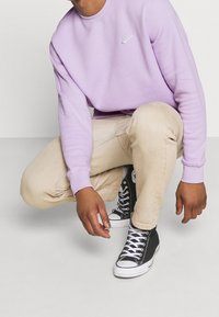 Ljuslila sweatshirt med rund hals och vit Nike-logotyp; beiga byxor; svarta höga sneakers med vit sula och snören. Avslappnad outfit.