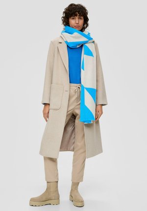 Beige langer Mantel, blauer Pullover, beige Hosen, blau-weißer gemusterter Schal, beige Stiefel mit dicken Sohlen, strukturierter Stoff.