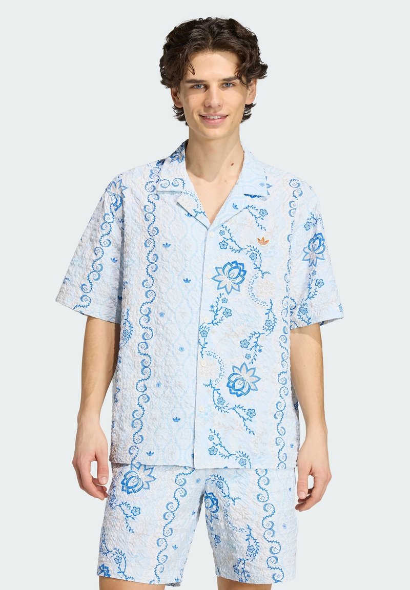 Jonge man staand, gekleed in een lichtblauw overhemd met korte mouwen en bijpassende korte broek met blauwe bloemen- en spiraalpatronen.